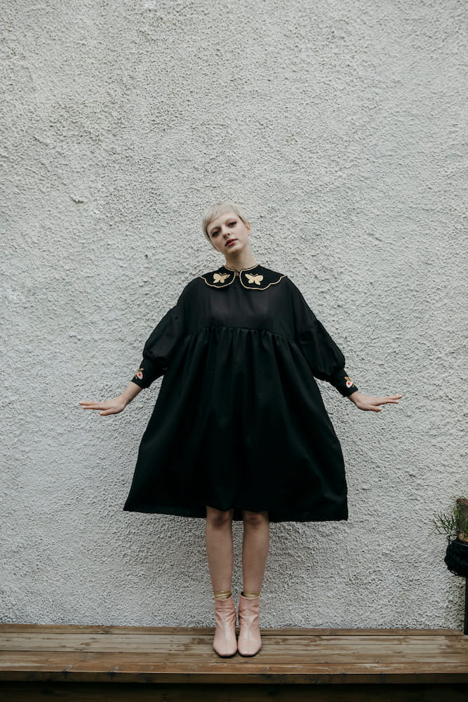 A person with short blond hair stands on a wooden platform against a textured white wall, wearing a loose black dress with an Estée Preda embroidery collar from the Noémiah Olivia Black Collar collection and pink boots. The person's arms are gently outstretched to the sides.