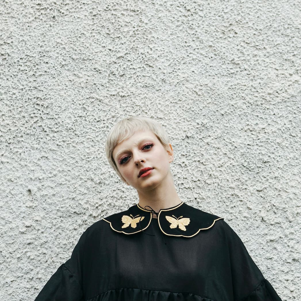 A person with short blonde hair stands against a textured white wall, wearing the Olivia Black Collar — Plain Back by Noémiah, adorned with gold butterfly designs. Their head is slightly tilted, and their eye makeup is strikingly artistic.