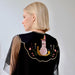 A woman with blonde hair is seen from the side wearing the oversized Olivia Black Collar by Noémiah, which features sheer sleeves. The back showcases an intricate embroidery of a snail with a house on its shell, adorned with flowers and floral patterns.