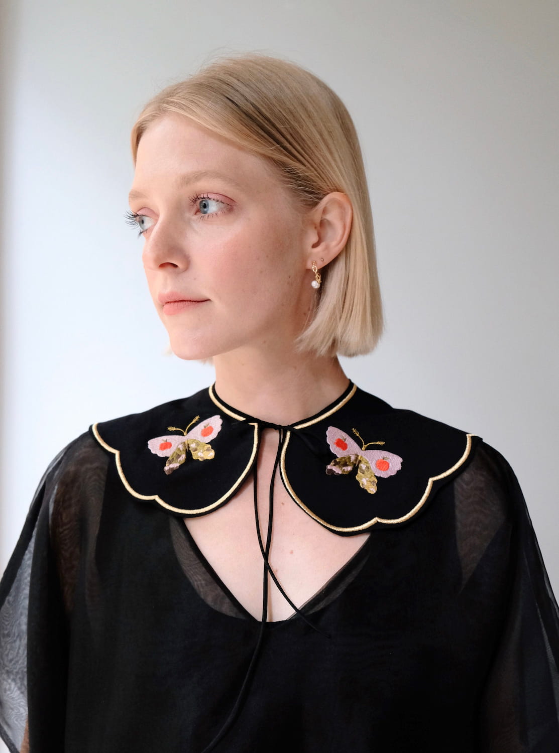 A person with short blonde hair is wearing the Olivia Black Collar top by Noémiah. It features sheer sleeves, an oversized Peter Pan collar adorned with embroidered butterflies and flowers. The person is looking to the side against a plain background.