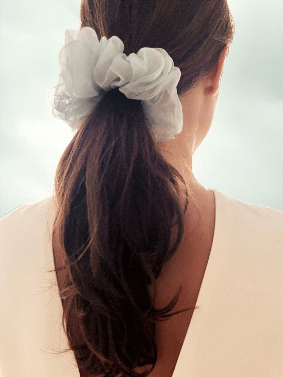 A woman with long brown hair is seen from behind, wearing a white top and a large scrunchie from Noémiah, in a light blue color. The background is softly blurred in light blue tones, evoking a serene ambiance.