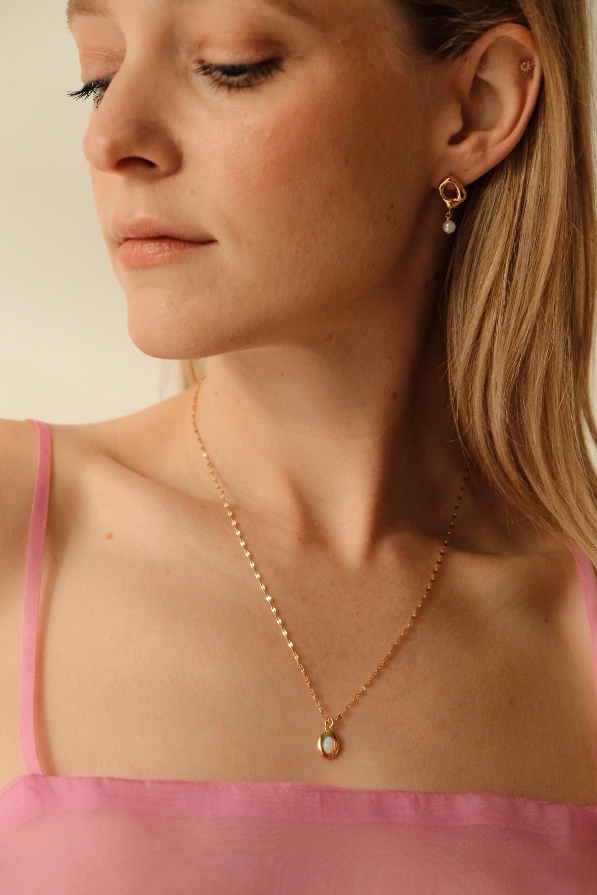 A woman with long blond hair is wearing a pink spaghetti strap top and gold jewelry. She has the Opaline Pendant by Noémiah on a delicate lace chain and gold-plated drop earrings. Her head is slightly turned to the side, displaying a calm expression.