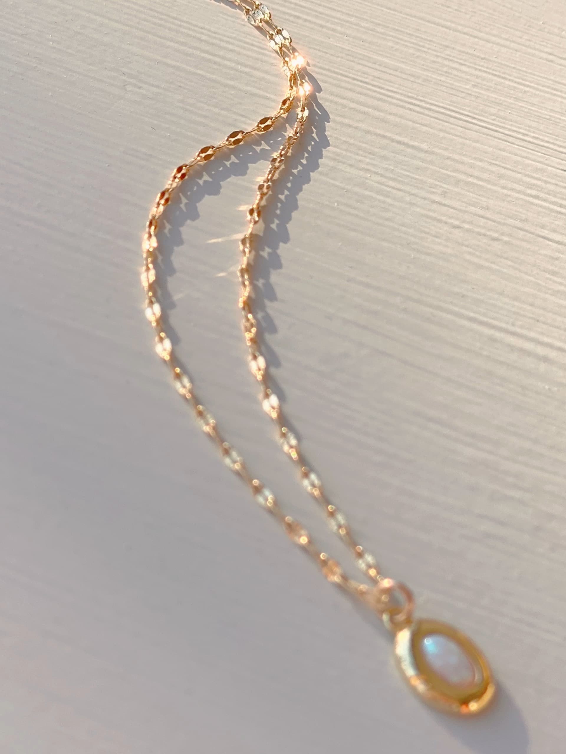 An Opaline Pendant from Noémiah, featuring a delicate lace chain necklace, rests on a light surface. The gold-plated, luminous iridescent stone emits a soft glow in the overall lighting.