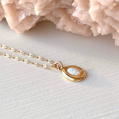 The Opaline Pendant by Noémiah, a gold-plated lace chain necklace featuring an oval opal pendant, elegantly rests on a light surface. The opal reveals subtle iridescent colors, beautifully complemented by pink crystalline formations in the background.