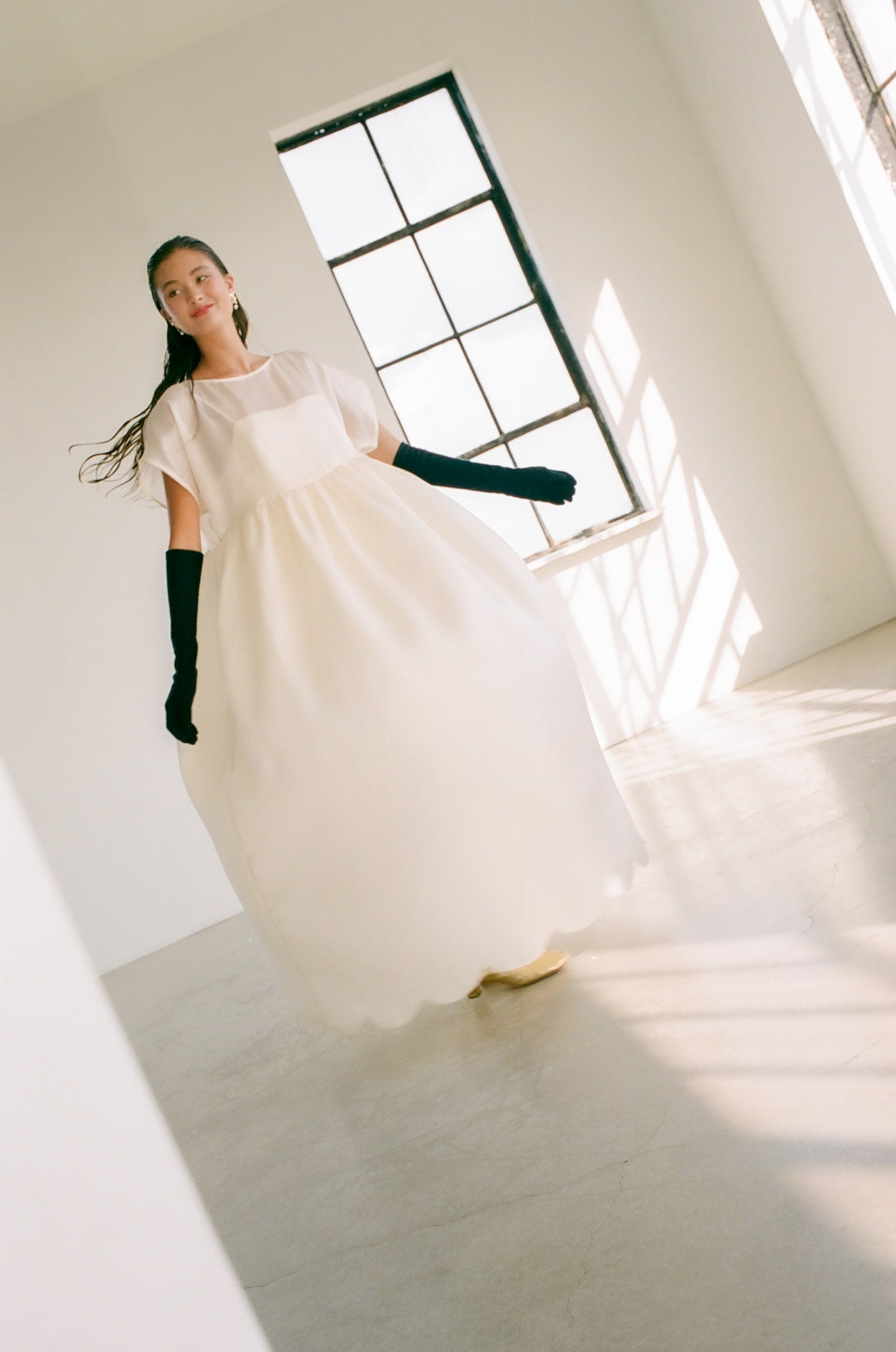 A woman in a flowing, white dress twirls gracefully in a sunlit room. Adorning her are Noémiah's Orchidée Earrings with freshwater natural pearls that glint softly in the light. Her long black gloves contrast elegantly, while the large windows cast gentle shadows on the floor.