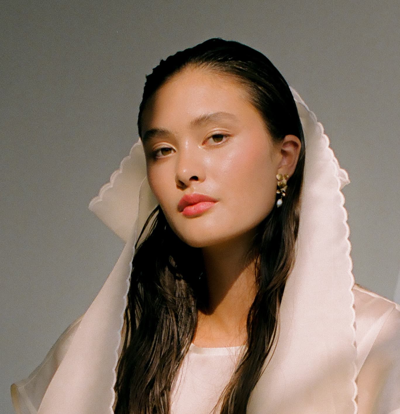 A woman with long dark hair gazes at the camera, wearing a sheer, scalloped-edged veil and the Orchidée Earrings by Noémiah, adorned with freshwater natural pearls. The lighting casts a soft glow on her face, highlighting her calm expression against a neutral background.