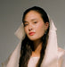 A woman with long dark hair gazes at the camera, wearing a sheer, scalloped-edged veil and the Orchidée Earrings by Noémiah, adorned with freshwater natural pearls. The lighting casts a soft glow on her face, highlighting her calm expression against a neutral background.