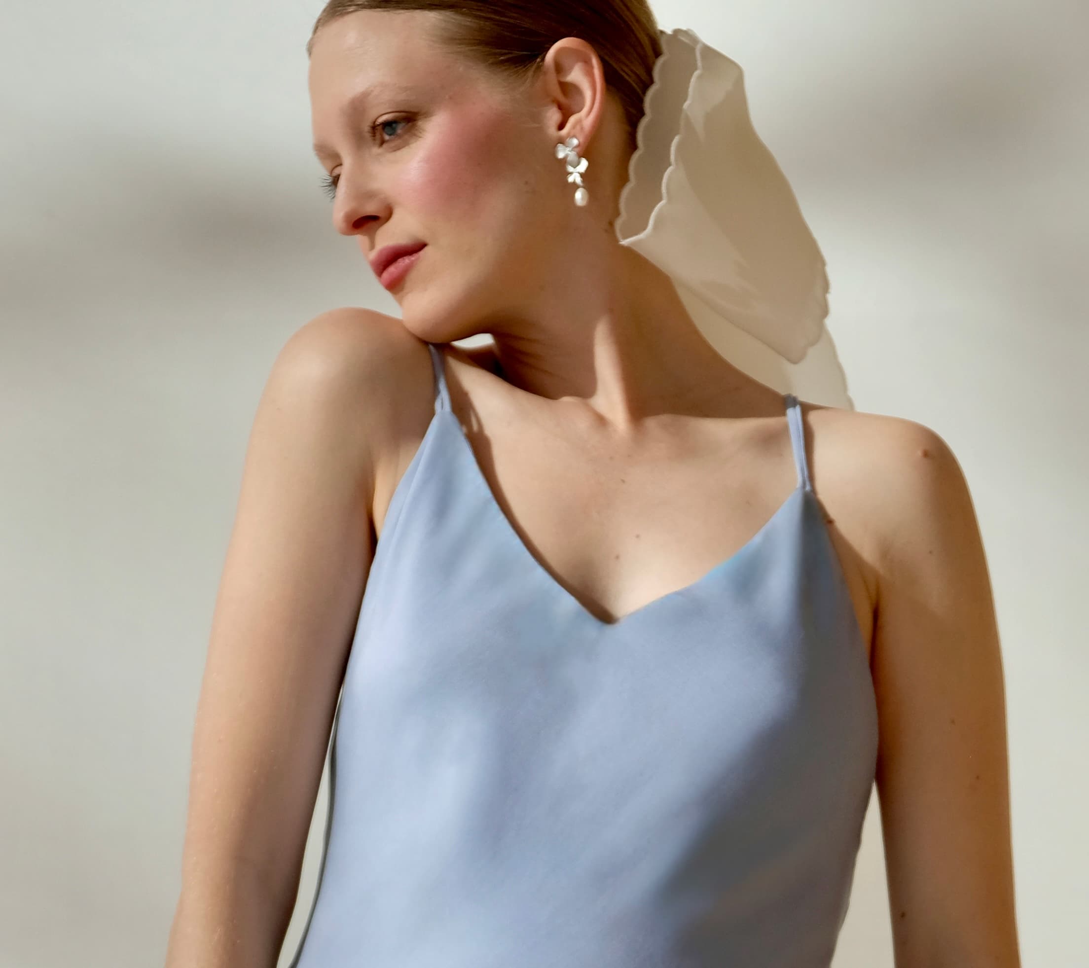 A person in a light blue sleeveless dress poses with their head turned to the side, wearing Noémiahs Orchidée Earrings adorned with freshwater natural pearls and a white fabric headpiece. The softly lit background casts delicate shadows.