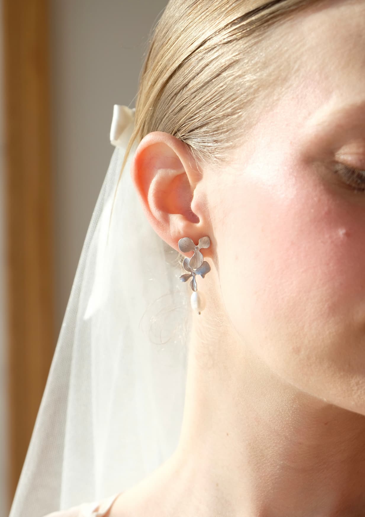 A close-up reveals a womans ear adorned with Noémiahs Orchidée Earrings, showcasing circular and floral designs accented by freshwater natural pearls. Her hair is tied back with a white bow, and a sheer veil drapes softly over her head, while soft light illuminates her skin.