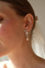 Close-up of a person wearing Noémiahs Orchidée Earrings with flower-shaped, tarnish-resistant plating. A white sheer veil is tied in their hair, capturing the lower half of their face against a softly lit background.