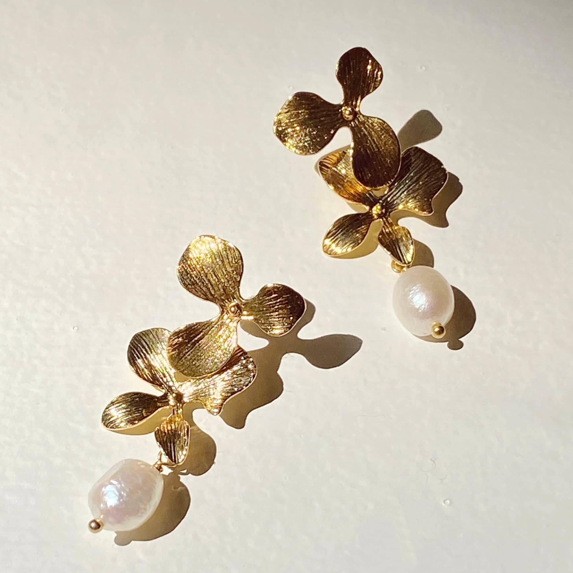 The Orchidée Earrings by Noémiah feature textured gold floral petals adorned with dangling freshwater natural pearls, all crafted with tarnish-resistant plating to ensure lasting elegance against a light background.