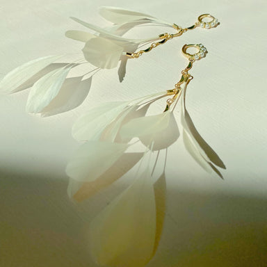 The Pamela Earrings by Noémiah are sophisticated, tarnish-resistant gold-plated earrings featuring delicate, translucent white petal-like shapes attached to gold hoops, elegantly resting on a light surface with gentle shadows.