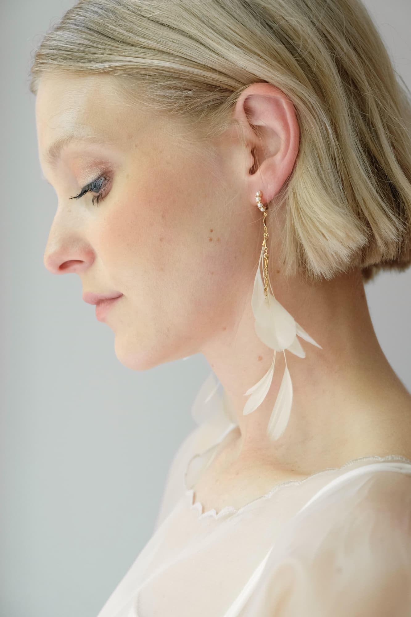 Profile of a person with short blonde hair wearing Noémiah's Pamela Earrings, which are delicate, tarnish-resistant gold-plated earrings shaped like leaves. The person is dressed in a sheer, light-colored top, and their eyes are gently closed against a soft background.