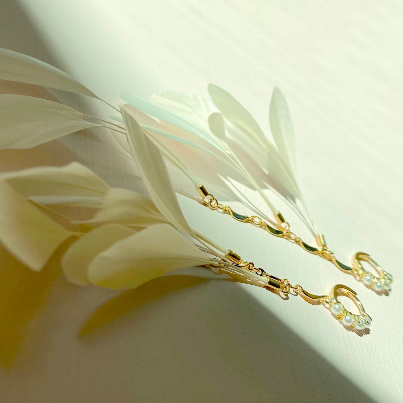 The Pamela Earrings by Noémiah, featuring delicate gold adorned with small pearls, rest gracefully on a white surface, set off by ivory feathers. Soft sunlight creates gentle shadows that highlight their tarnish-resistant gold-plating, resulting in a serene and elegant composition.