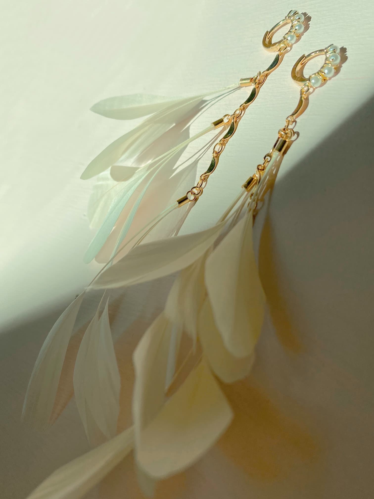 The Pamela Earrings by Noémiah are dainty gold-plated accessories, designed to be tarnish-resistant and adorned with translucent ivory feather-like pendants that gently cast soft shadows on a light surface.