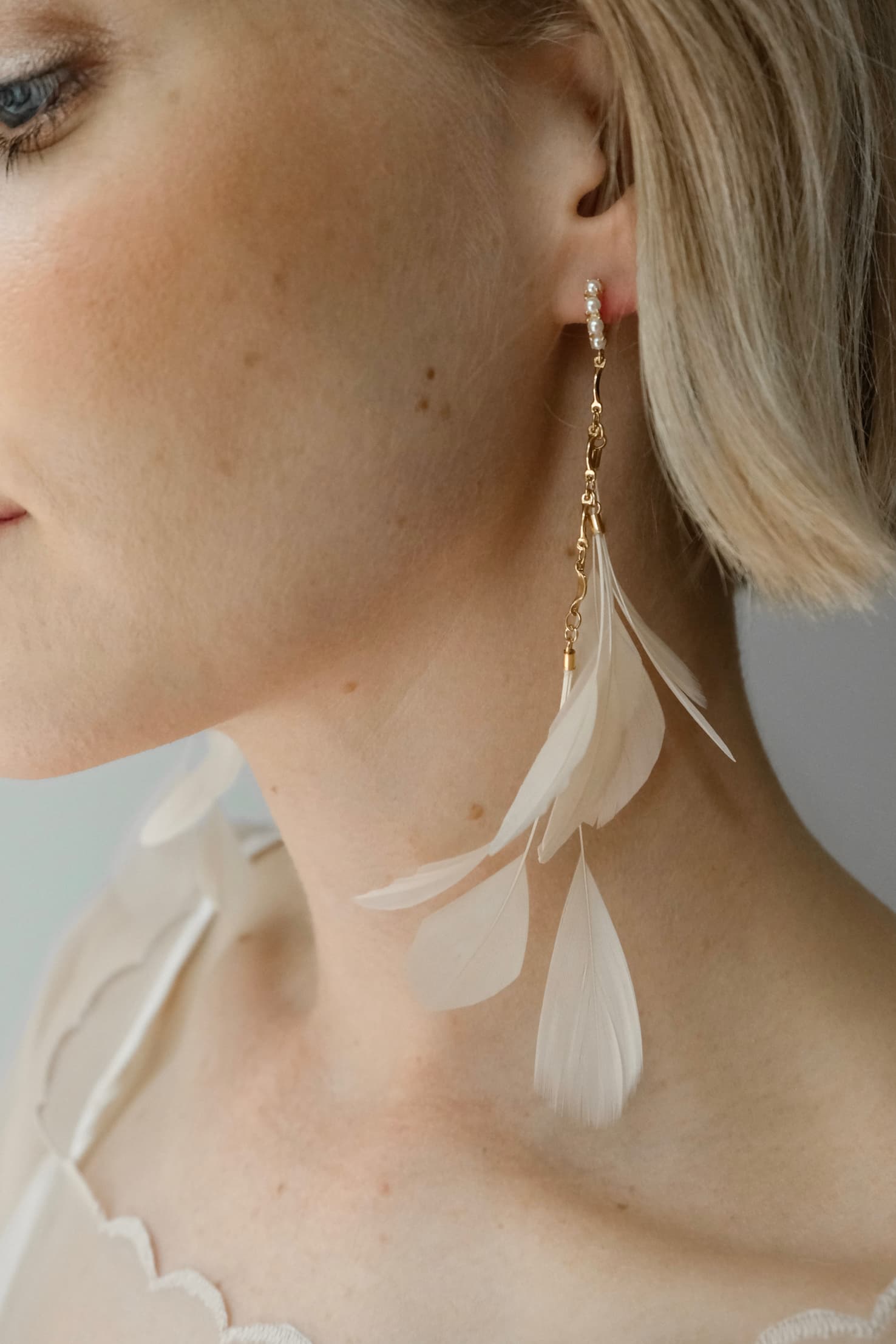 A close-up of a woman showcasing the Pamela Earrings by Noémiah, which feature delicate, tarnish-resistant gold plating and are adorned with ivory feathers. She has short blonde hair and is dressed in a light-colored top with scalloped edges, set against a soft and neutral background.