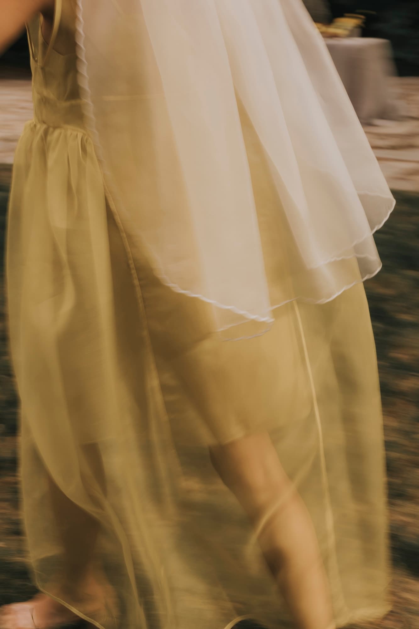 A person wearing the Paris Veil, a flowing translucent yellow dress from Noémiah, moves gracefully outdoors. The embroidered edges enhance the blurred motion of the silk organza fabric, imbuing the image with an ethereal quality through its soft focus.