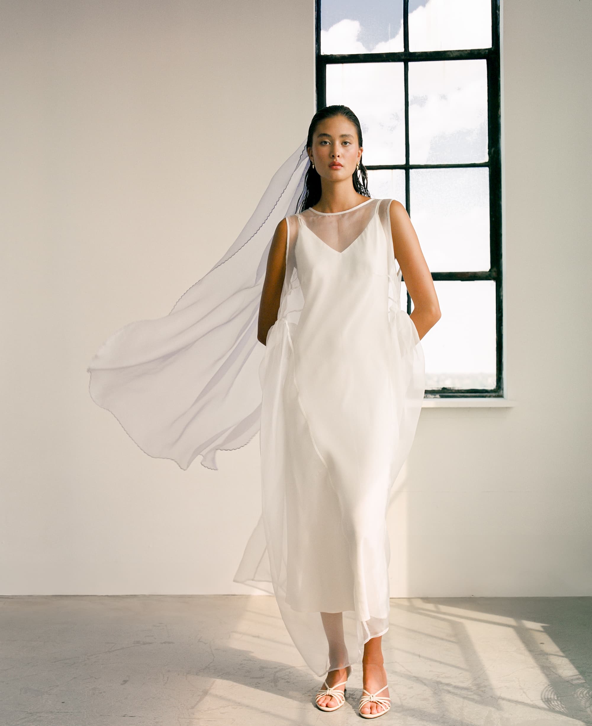 A woman stands in a minimalist room, her flowing white silk organza dress and the matching Paris Veil by Noémiah with embroidered edges catching the sunlight streaming through an open window behind her onto the floor. Her hair is slicked back, enhancing the elegant simplicity of her look.