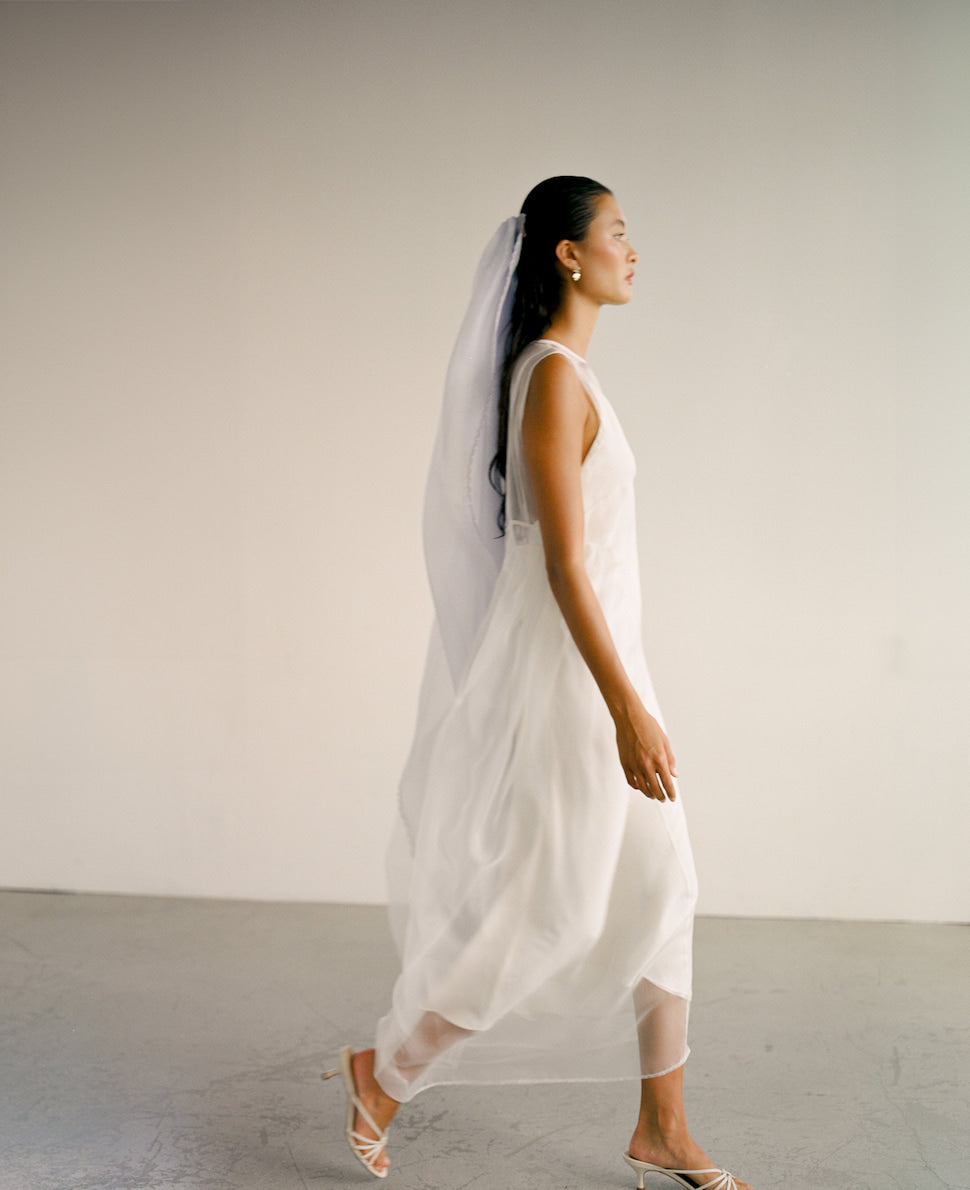 Wearing a flowing white dress and the exquisite Paris Veil by Noémiah, a woman gracefully walks across a plain background. She is donning open-toed heels, and her long hair is pulled back. The minimalist scene highlights her elegant attire and the intricate embroidered edges of the veil.