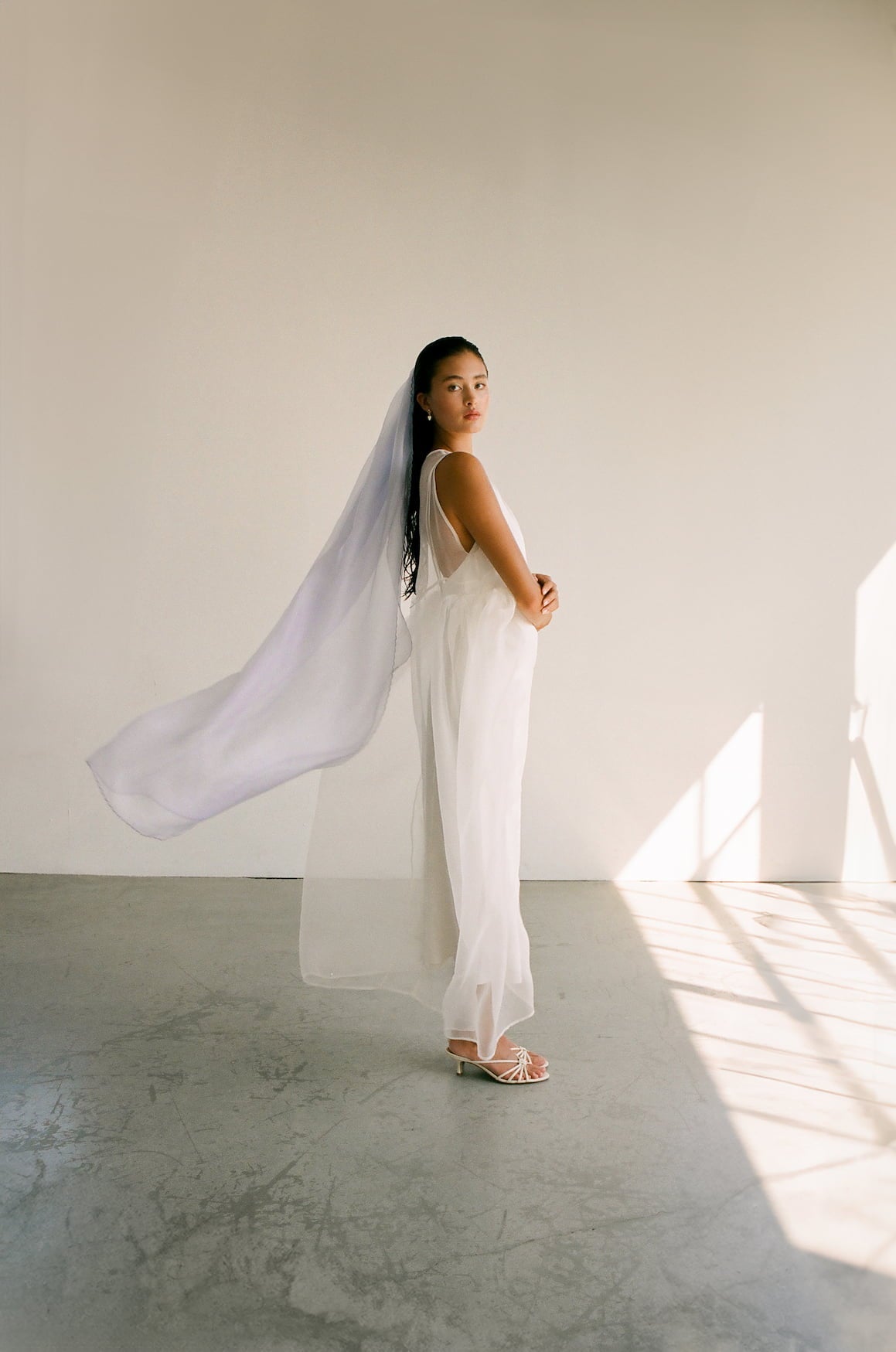 In a minimalist room with soft lighting, a woman stands wearing the Paris Veil by Noémiah, which features embroidered edges that trail gracefully behind her. She is also dressed in a flowing white dress and faces sideways towards the camera, with her hands clasped gently in front.