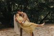 A groom in a beige suit joyfully lifts and embraces his bride, who is wearing a yellow dress and a Noémiah Paris Veil with embroidered edges. They are outdoors on a stone path, surrounded by lush green trees. The scene is filled with happiness and romance.