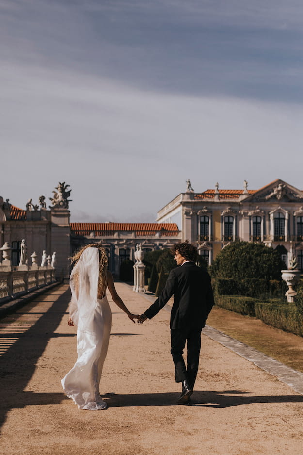 A bride and groom stroll hand in hand along a sunlit path with a historic building in the background. The bride's long white dress flows elegantly, featuring silk organza and embroidered edges, while her Paris Veil by Noémiah catches the light. The sky is clear and blue, surrounded by well-manicured gardens.