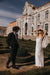 A couple in wedding attire stands in a formal garden with manicured hedges. The bride, wearing a white silk organza gown and the Paris Veil by Noémiah, shields her eyes from the sun. The groom is in a dark suit. An ornate building with intricate details is in the background.