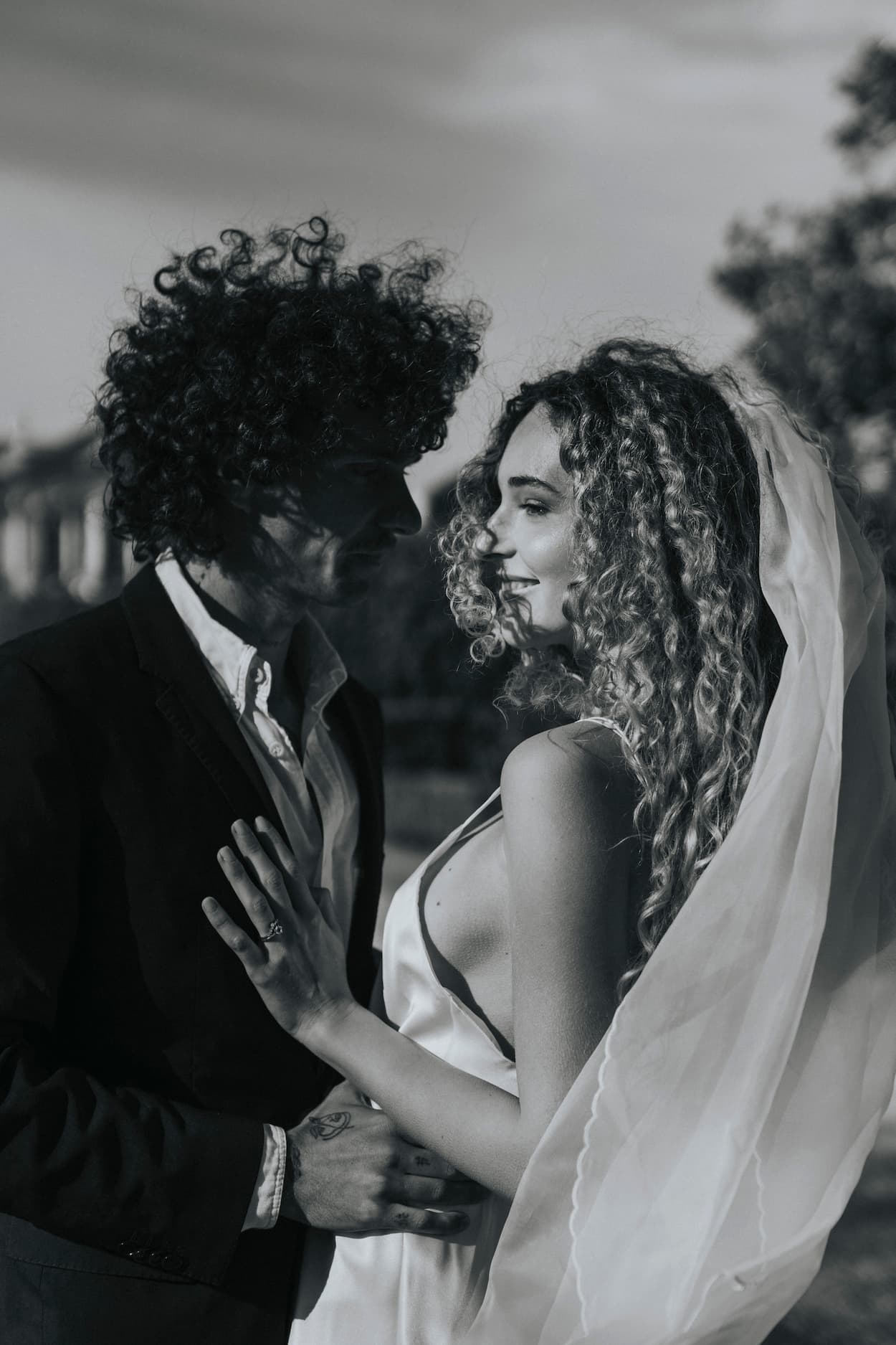 A black and white photo captures a couple on their wedding day. The bride, in a silk organza dress with embroidered edges, faces the groom with his curly hair and suit. Her Paris Veil by Noémiah catches the light as they gaze into each other's eyes, sharing a tender moment outdoors.