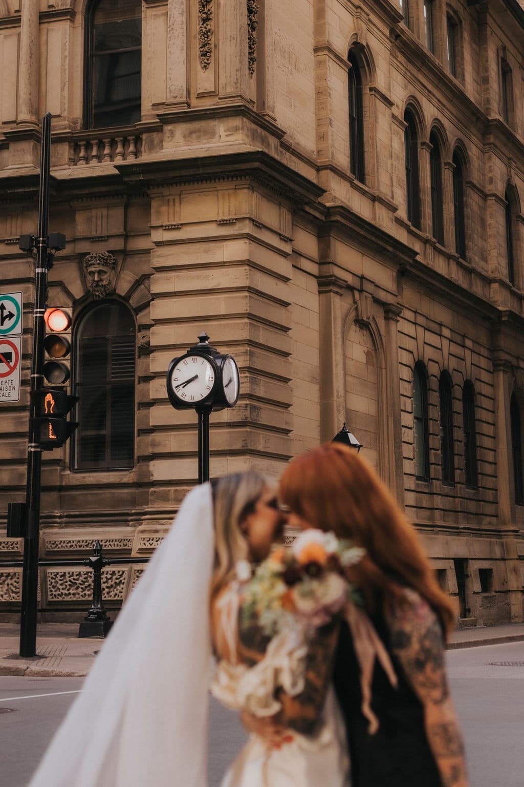 Two people embrace, partially hiding their faces behind a bouquet of flowers. The delicate silk organza of a Noémiah Paris Veil drapes elegantly, as they stand before an ornate, historic building with a street clock and traffic lights in the background.