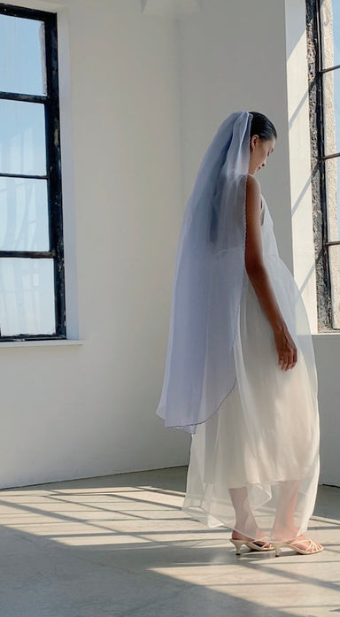 A woman in a flowing white dress and the Paris Veil by Noémiah stands in a sunlit room with large windows. The veil’s embroidered edges catch the light, while she appears contemplative, with silk organza fabric creating soft shadows on the floor.