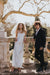 A woman in a white dress with embroidered edges and wearing the Paris Veil by Noémiah stands next to a man in a black suit. They are leaning against a stone balustrade, surrounded by trees, suggesting an outdoor, formal setting.