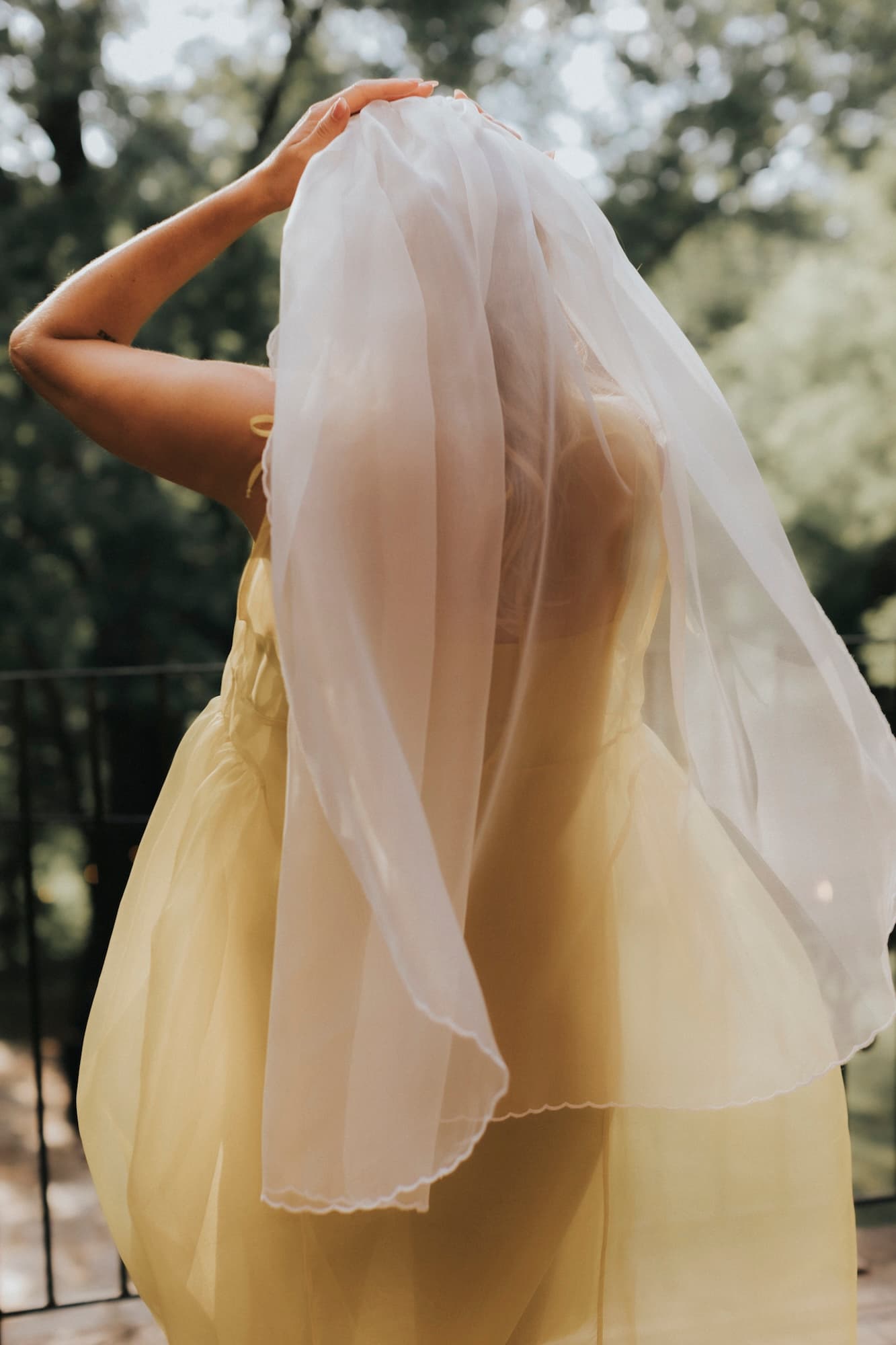 A person wearing a light, translucent yellow dress made of silk organza and a white Paris Veil by Noémiah, featuring embroidered edges, stands on a balcony facing away. The background showcases blurred green trees, hinting at a peaceful outdoor setting.