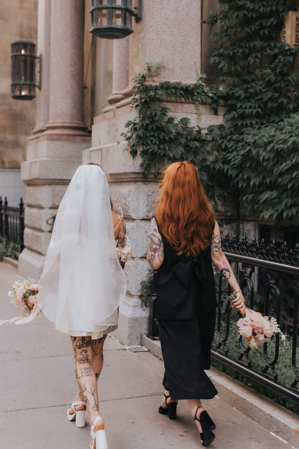 A bride in a white silk organza dress and the Paris Veil by Noémiah walks alongside a woman in a black dress. Both are holding bouquets, with the bride displaying tattoos. They stroll down a city sidewalk, surrounded by ivy-covered stone buildings and vintage-style lanterns.