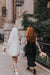 A bride in a white silk organza dress and the Paris Veil by Noémiah walks alongside a woman in a black dress. Both are holding bouquets, with the bride displaying tattoos. They stroll down a city sidewalk, surrounded by ivy-covered stone buildings and vintage-style lanterns.