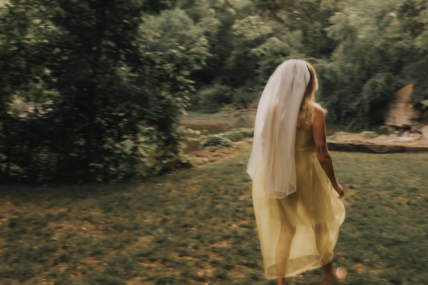 A woman in a pale yellow dress and a Paris Veil by Noémiah, adorned with embroidered edges, walks on a grassy area surrounded by lush green trees. Her back faces the camera, adding to the scene's soft, blurred effect that creates a dreamy atmosphere.