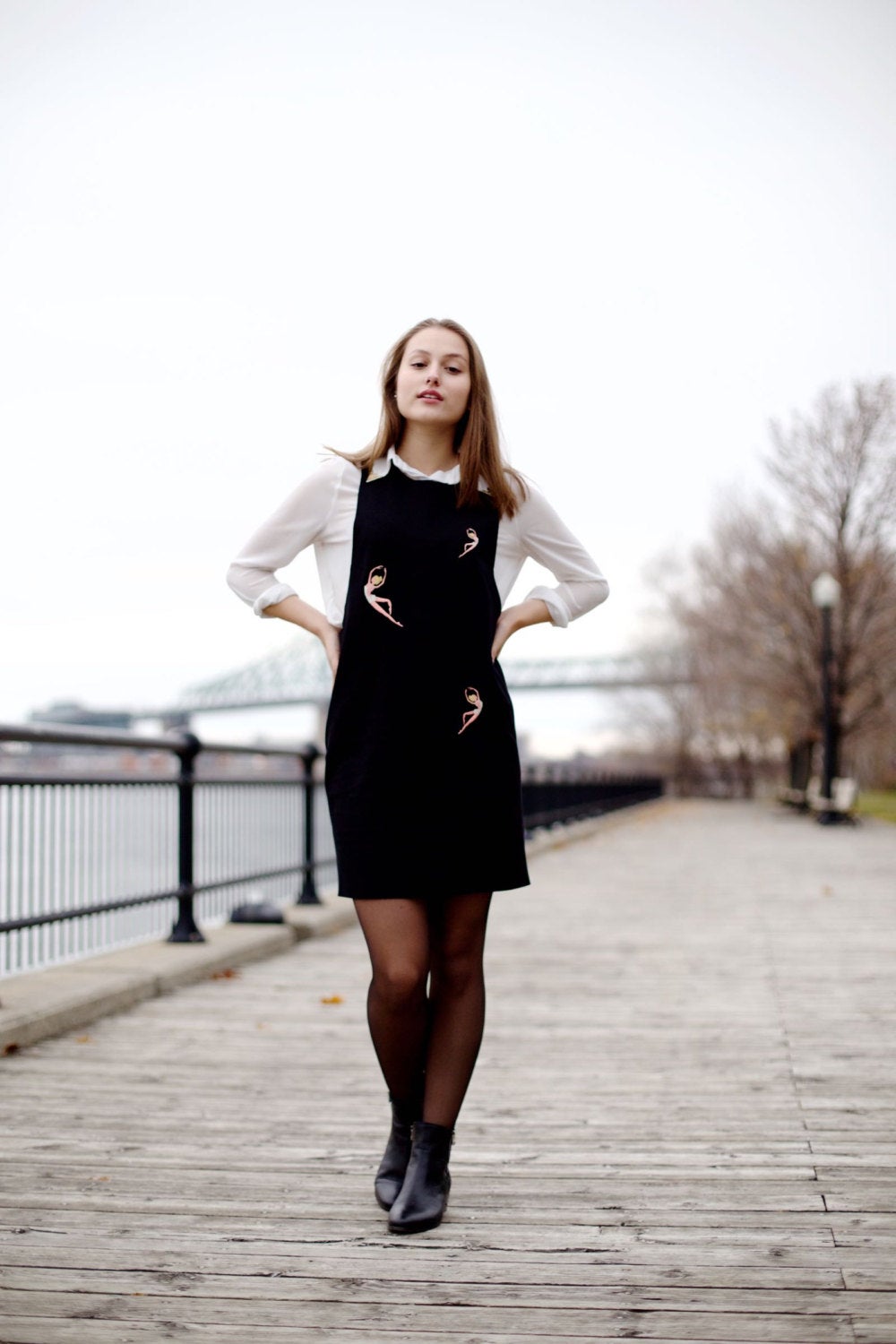 A woman stands on a wooden boardwalk beside a river, hands on her hips. She is wearing the Paule Dress by Noémiah, elegantly layered over a white shirt and paired with black ankle boots. In the background, a bare tree and bridge under a cloudy sky enhance her stylish ensemble.