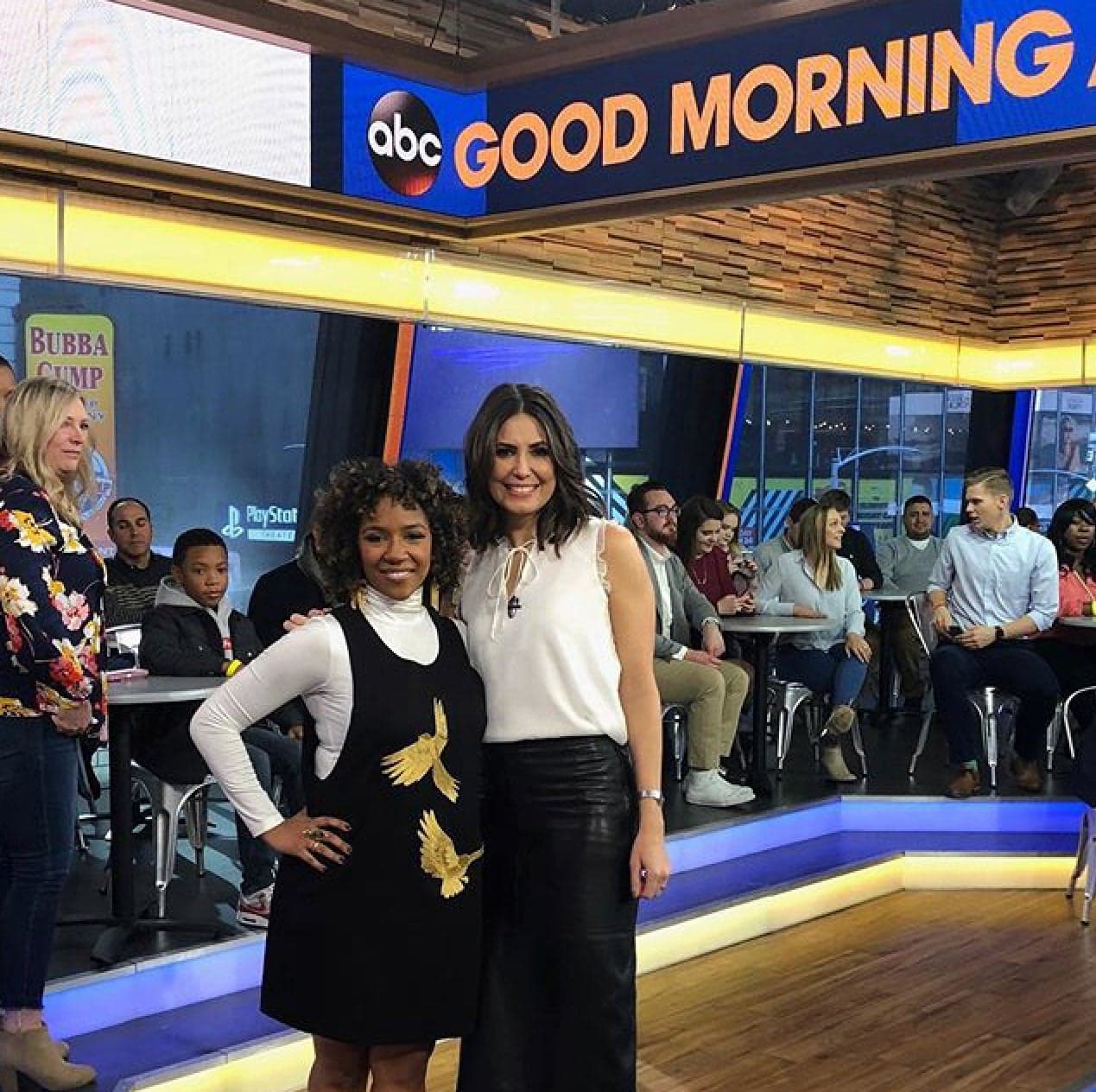 Two women, dressed in the Paule Dress by Noémiah, stand together, smiling, on a TV show set with "Good Morning" displayed above. They are surrounded by an audience seated at tables. The studio is adorned with custom fit designs and embroidered fabric accents, featuring large windows and bright lights.