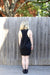 A person with shoulder-length blonde hair stands facing a wooden fence, wearing the Paule Dress by Noémiah, a sleeveless black dress crafted from double-knit fabric, paired with black shoes. In the background, a tree with green leaves and a partially visible brick wall above the fence can be seen.