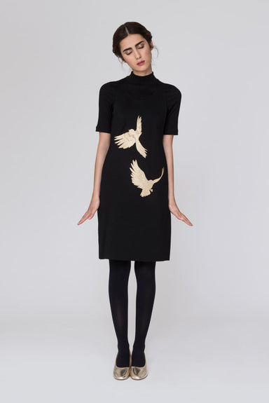 A woman with closed eyes stands wearing the Paule Dress by Noémiah, crafted from double knit fabric and adorned with two gold bird silhouettes. She pairs it with black leggings and custom-fit gold shoes against a plain gray background. Her arms rest by her sides, and she leans slightly forward.