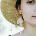 A close-up of a person wearing a straw hat highlights the right side of their face, elegantly adorned with Pénélope Earrings from Noémiah, crafted with high-quality feathers. The softly blurred green background adds to the serene ambiance.