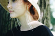 A woman wearing a wide-brimmed hat stands in soft, dappled sunlight. Her long, high-quality Pénélope Earrings from Noémiah complement her black top beautifully. The background, with its blurred tree trunks and greenery, adds to the serene ambiance.