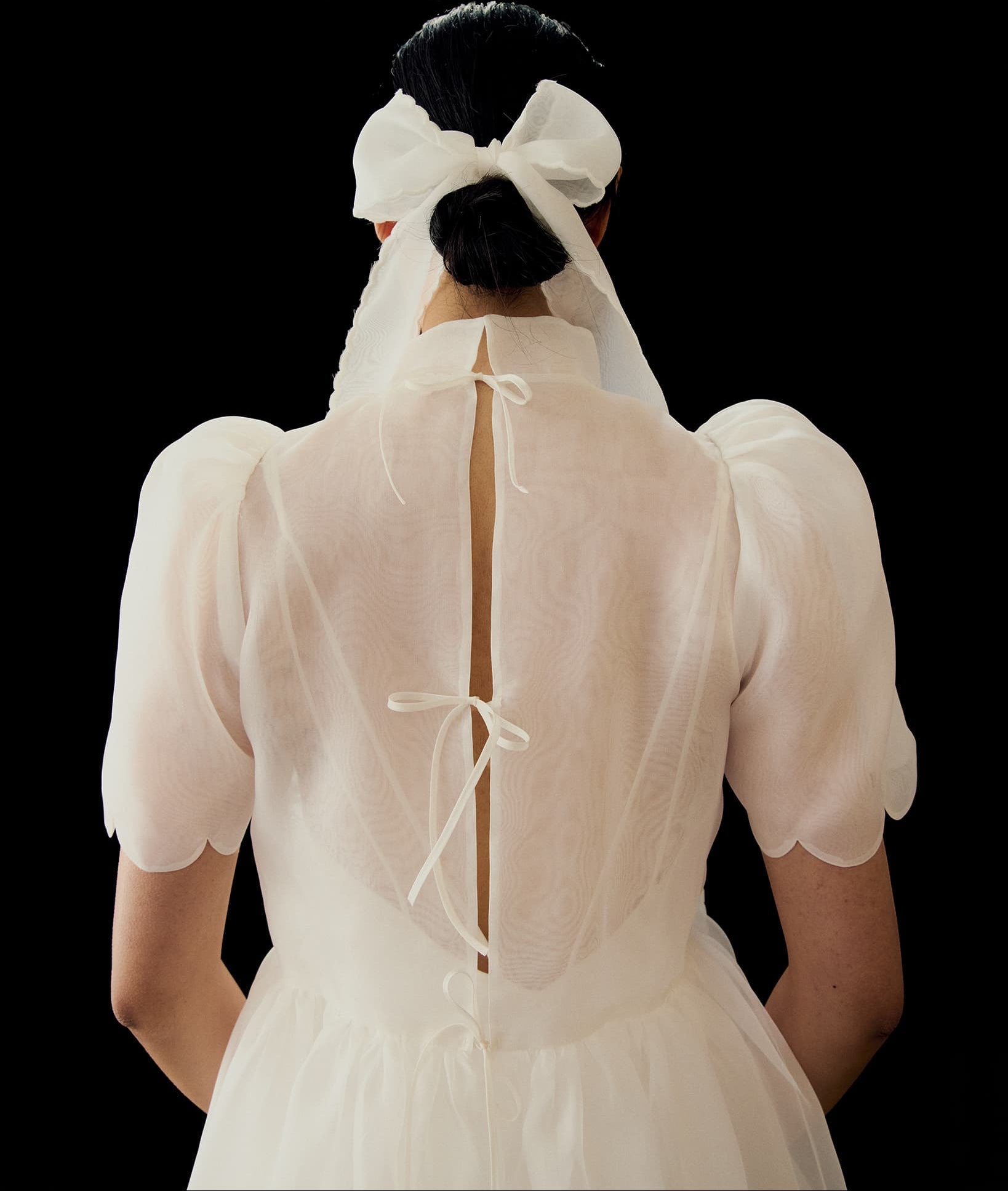 A person in a sheer white dress with puffed sleeves and scalloped edges is seen from behind, their hair tied back with the Noémiah Silk Ribbon — Classic against a black background.