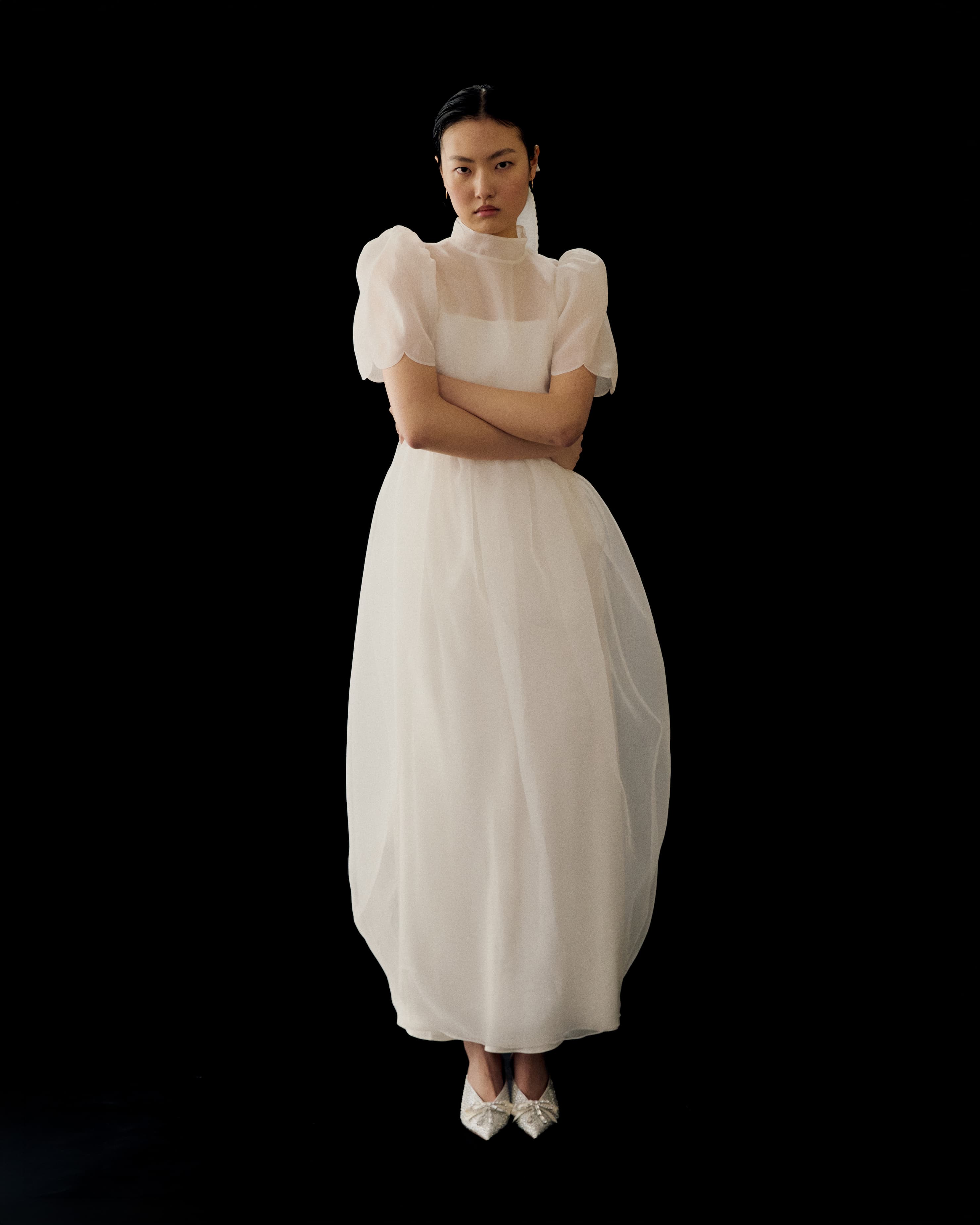 A woman stands against a black background wearing Noémiah’s Perle Dress — Bridal Edit, a long sheer silk organza gown with puffed short sleeves, paired with white pointed shoes. Her arms are crossed and her expression is neutral.