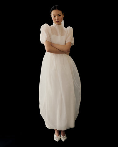 A woman stands against a black background wearing Noémiah’s Perle Dress — Bridal Edit, a long sheer silk organza gown with puffed short sleeves, paired with white pointed shoes. Her arms are crossed and her expression is neutral.