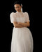 A woman stands against a black background, wearing the Noémiah Perle Dress — Bridal Edit, a sheer white silk organza design with puffed short sleeves. Her arms are crossed and her dark hair is tied back with a white ribbon as she gazes down thoughtfully.