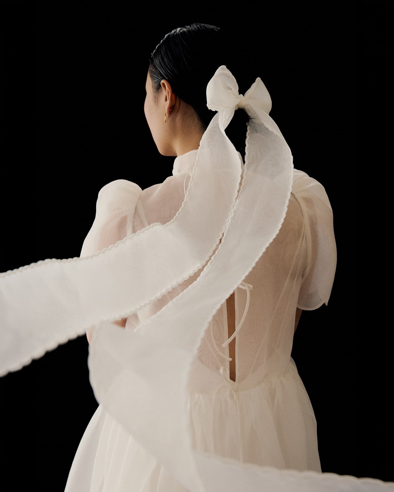 A person in the Noémiah Perle Dress — Bridal Edit, featuring sheer silk organza and puffed sleeves, stands with their back to the camera. Their dark hair is tied with a large white ribbon flowing outward against a solid black background.