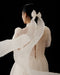 A person in the Noémiah Perle Dress — Bridal Edit, featuring sheer silk organza and puffed sleeves, stands with their back to the camera. Their dark hair is tied with a large white ribbon flowing outward against a solid black background.