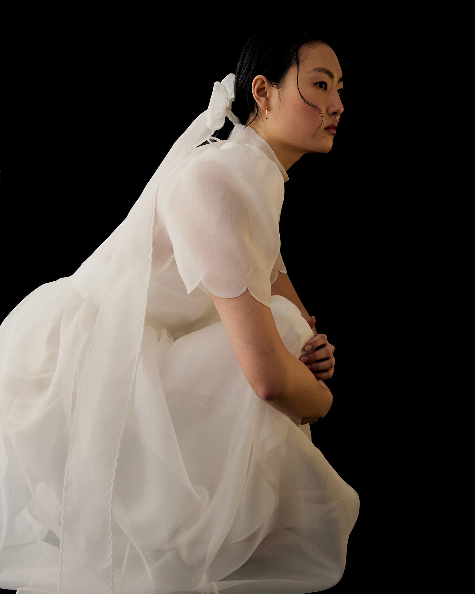 A person in the Perle Dress — Bridal Edit by Noémiah, featuring sheer silk organza and puff sleeves, crouches against a black background with a long ribbon in their hair, gazing to the side.