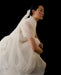 A person in the Perle Dress — Bridal Edit by Noémiah, featuring sheer silk organza and puff sleeves, crouches against a black background with a long ribbon in their hair, gazing to the side.