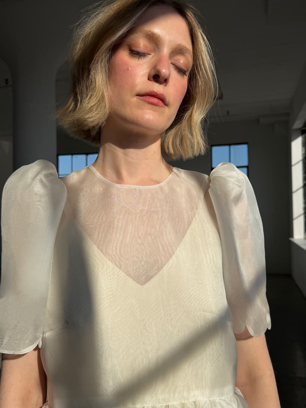 A woman with short blonde hair stands in soft sunlight, eyes closed, wearing the Noémiah Claude Silk Dress — Bridal Edit. Gentle shadows fall across her as large windows frame the minimal room behind her.